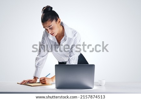 Job and office work concept with young pretty businesswoman in white classic shirt making notes in diary leaning on white wooden table with modern computer and coffee cup on light grey wall background