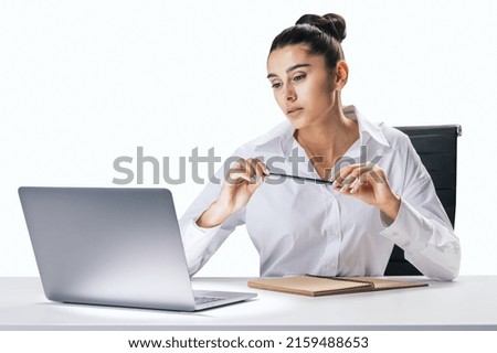 Business process and job concept with pretty young woman sitting at white desk, looking at laptop screen and carrying pencil in hands on abstract white wall background