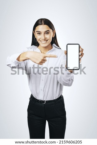 Mobile application concept with smiling businesswoman in white shirt showing modern cellphone with blank white screen on abstract light grey background, mockup