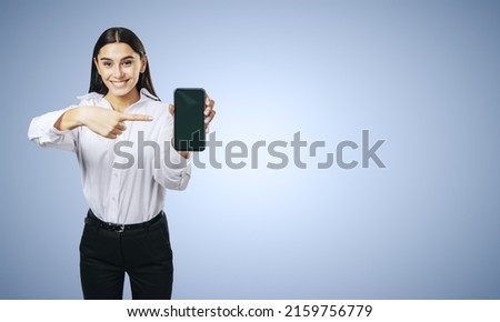 Mobile application concept with happy girl in white shirt showing modern smartphone with blank screen on abstract light blue background with place for your logo or text, mockup