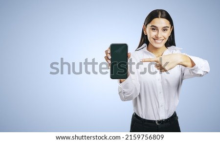 Mobile application concept with smiling businesswoman in white shirt showing modern smartphone with blank screen on abstract light blue background with place for your logo or text, mock up