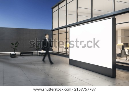 Side view businessman passing by outdoor blank white billboard and stylish street benches near modern business center building with glass walls in the evening