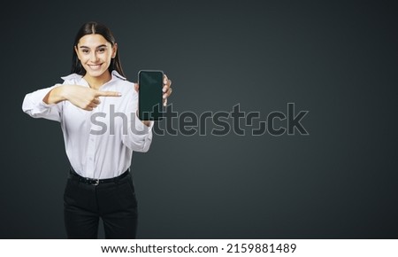 Mobile application concept with smiling businesswoman in white shirt showing modern smartphone with blank screen on abstract black background with place for your logo or text, mockup