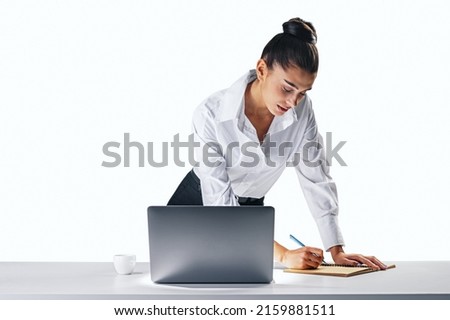 Job and office work concept with young pretty businesswoman in white classic shirt making notes in her diary leaning on white wooden table with laptop and coffee cup on light wall background