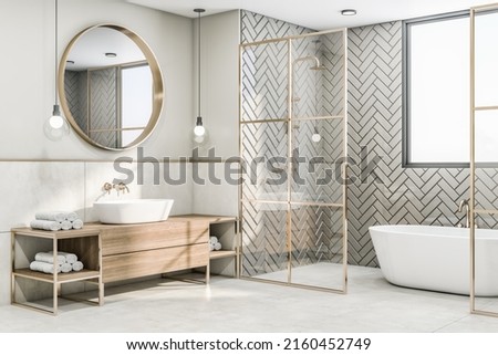 Perspective view on sunny modern interior design bathroom with herringbone ceramic tales bath zone, wooden sink cabinet under round mirror and glass partitions with gold decorations. 3D rendering