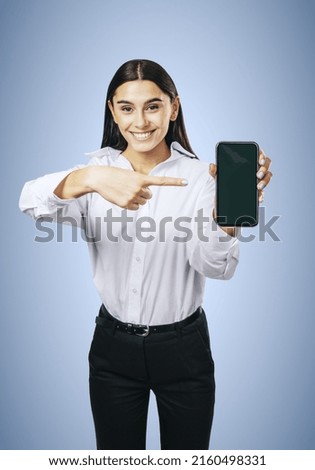 Mobile application concept with happy girl in white shirt showing modern smartphone with blank screen on abstract light blue background, mockup