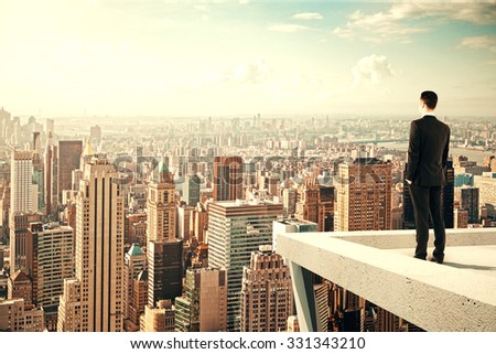 Empresario parado en el techo de un rascacielos y mirando a la ciudad al atardecer