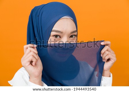 Asian young Muslim woman holding hijab headscarf covering her face. Muslim woman hiding her face behind hijab headscarf on orange colour background in studio