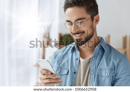 Happy attractive male European model looks happily at mobile phone, reads positive news from newsfeed, sends message to lover during work break. Young man recieves email on modern smart phone