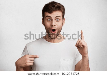 Amazed male model with beard raises fore finger as gets some brilliant idea, dressed in casual white t shirt, indiacates, isolated over white concrete wall. Handsome surprised man gestures indoor