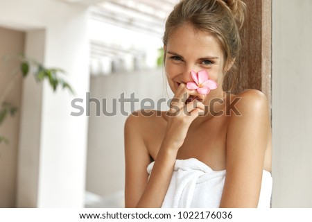 People, resort and recreation concept. Good looking natural shy young woman rests in tropial hotel, has honey moon with husband, smells orchid flower, wrapped in bath towel, takes shower or bath