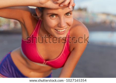 Close up shot of cheerful female jogger tired from morning workout, being sweaty, being on coastline of tropical island, has positive expression. People, active lifestyle, outdoor sport concept