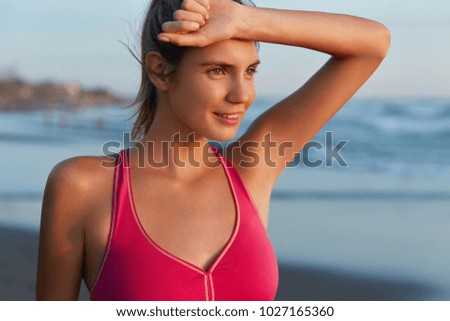 Outdoor shot of tired female athlete wears sports clothes, being sweaty, going to swim in sea, looks tired but satisfied with cardio training on coastline. Fatigue jogger wins sport competition