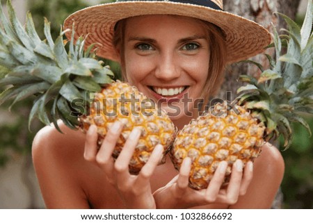 Horizontal shot of happy attractive female with broad smile, holds two pineapples, enjoys rest in tropical villa, eats exotic fruits or going to make cocktail. Female tourists tastes juicy fruit 商業照片 © 