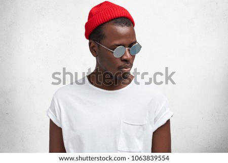 Isolated shot of fashionable serious young African American male in sunglasses, red trendy hat and casual t shirt, looks aside, isolated over white studio background. People and style concept