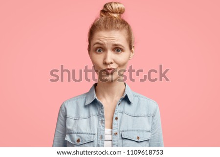 Unhappy blonde European female with discontent expression, frowns face, recieves bad news from interlocutor, stands against pink background. Disappointed young woman in denim jacket has no make up