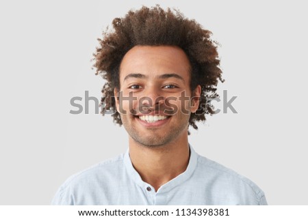 Mixed race curly male with broad smile, shows perfect teeth, being amused by interesting talk, has bushy curly dark hair stands indoor against white blank wall. Close up of joyful African American man