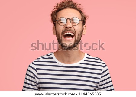 Overjoyed guy laughs joyfully as hears something funny, can`t stop happiness, has carefree lifestyle, wears spectacles, balck and white striped sweater, stands over pink background. Positive emotions