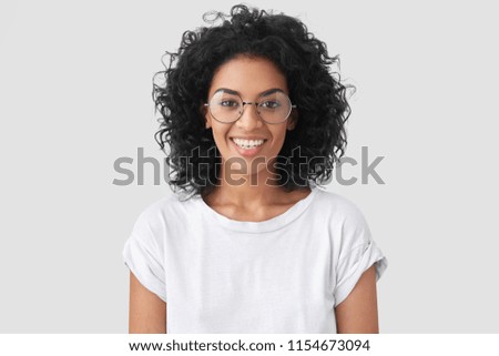 Beautiful smiling African American female with crisp hair, broad smile, shows white teeth, wears casual t shirt and spectacles, stands over studio wall rejoices having day off. Woman journalist indoor