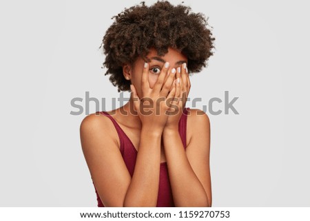 African American beautiful young female peeks though fingers, covers face with both hands, has frightened expression as notices something terrible or scarying, isolated over white background.