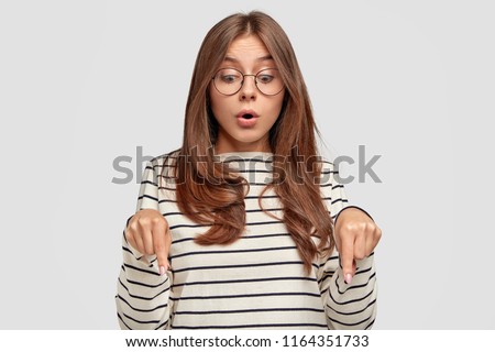 Surprised young lovely female teenager opens mouth widely, has startled look, points down with stupefied expression, sees something unbelievable, isolated over white background. Omg, look there!