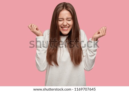 Successful brunette girl closes eyes from happiness, keeps raised fists, dressed casually, celebrates her victory with triumph, isolated over pink background. People, joy, body language concept