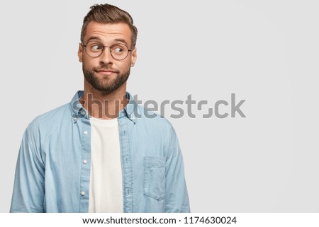 Isolated shot of funny bearded man designer or freelancer looks thoughtfully aside, thinks on how climb career ladder, wears casual shirt and round spectacles, isolated on white wall with copy space