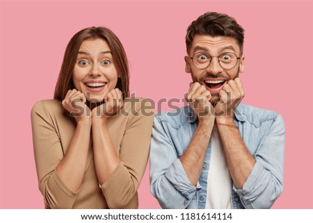 Shot of glad young brother and sister keep hands under chin, have broad smiles, rejoice good news, dressed in casual clothes, isolated over pink background, express happiness. Wow, thats great!