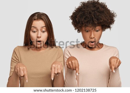 Photo of stupefied mixed race young Caucasian women point at floor together, have surprised gaze down, dressed casually, stand next to each other against white background. Wow, just look there!