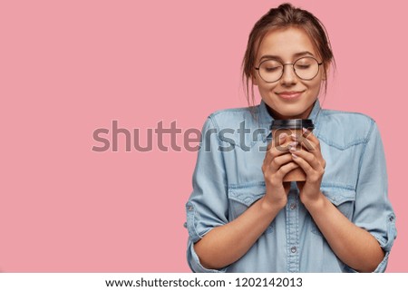 Pleased lovely Caucasian woman holds aromatic beverage, drinks cappuccino or coffee, feels warm, closes eyes from pleasure, has charming smile, wears denim shirt, isolated over pink background