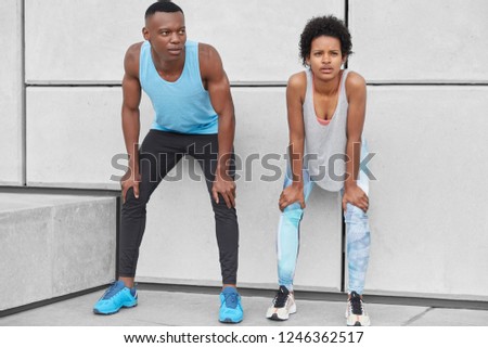Horizontal view of fatigue woman and man lean at knees, stand closely to white wall, catch breath after intensive run, wear sneakers, leggings and t shirt, have contemplative expressions at camera
