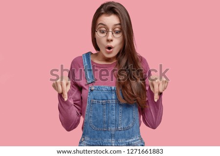 Studio shot of surprised Caucasian lady points with both index fingers on floor, being amazed and stunned, disappointed by something, isolated over pink bacckground. Omg, look at my dirty shoes