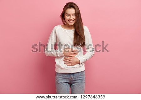 Satisfied smiling young woman keeps both hands on belly, being in good mood after eating delicious supper, demonstrates she is full, isolated over pink background. Pleasant feeling in stomach