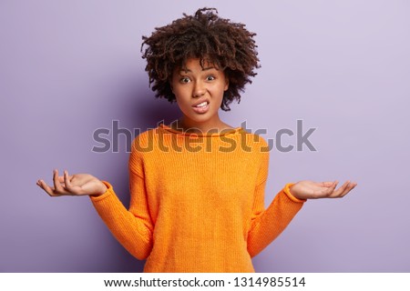 Isolated shot of confused beautiful woman with dark skin, curly haircut, spreads hands sideways, smirks face, feels doubt while makes choice, dressed in casual jumper, isolated over purple wall Foto stock © 