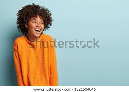 Glad overemotive dark skinned lady with curly hairstyle, laughs happily, expresses sincere emotions, being amused by friend, dressed in orange casual jumper, models in studio alone with mockup space