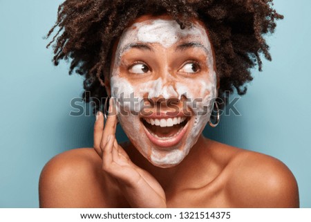Headshot of happy black woman with foam or foaming cleanser on face, has facial treatments, gazes aside, opens mouth, has Afro hairstyle, isolated over light blue wall. Perfect clean skin concept