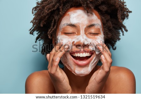 Spa, therapy and beauty concept. Satisfied black woman washes face with white soap, has cleansing foam, purification of face, cleans pores, shows bare shoulders, isolated over blue studio wall