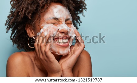 Sideways shot of pleasant looking lady applies cosmetic foam, feels pleasure after beauty treatments, polishes face, isolated over blue background with empty space on left side for your text