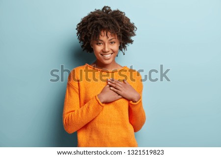 Touched beautiful smiling Afro American girl keeps palms on heart, expresses appreciation, pleasure and gratitude, wears orange sweater, models over blue background. Confession in love concept.