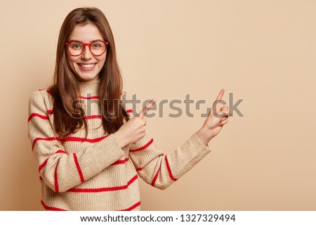 Horizontal shot of cheerful woman points away with both fore fingers, looks with admiration and interest at free space, demonstrates interesting cute product, dressed casually, isolated on beige wall