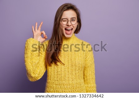 Waist up shot of pleased young woman says good job or well done, makes okay gesture, demonstrates symbol of approval and like, blinks eye, wears spectacles, knitted yellow jumper, stands indoor