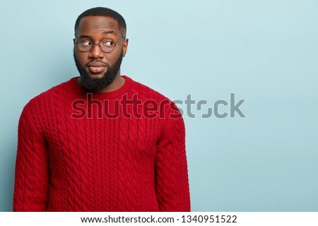 Suspicious guy looks away, suspects someone tells lie, thinks doubtful, has thick bristle, wears round spectacles and red outfit, isolated over blue studio wall with empty space for your advert