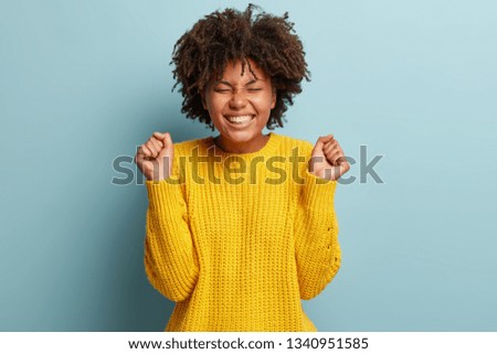 Overemotive triumphing woman squints face from excitement, clenches fists in victory, hears amazing good news, shows broad smile, wears oversized yellow jumper, gestures joyfully. Success concept