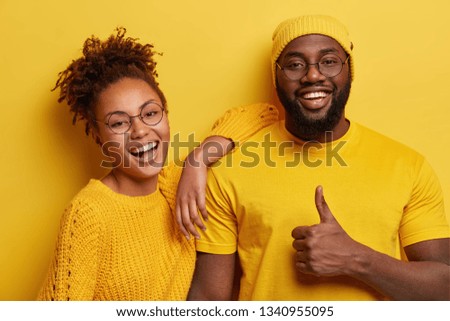 Horizontal shot of satisfied dark skinned woman laughs happily, leans at shoulder of boyfriend, come together on theme party, dressed in yellow clothes. Pleased bearded Afro man shows thumb up