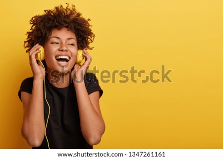 Happy young Afro American lady relaxed with great song, has broad smile, touches headphones, listens music, dressed in black casual t shirt, isolated over yellow studio wall, wants to dance.