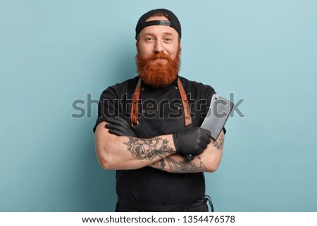 Portrait of handsome professional male butcher has arms crossed, holds metal sharp cleaver, wear black uniform, ready to start work, isolated over blue background. People and occupation concept