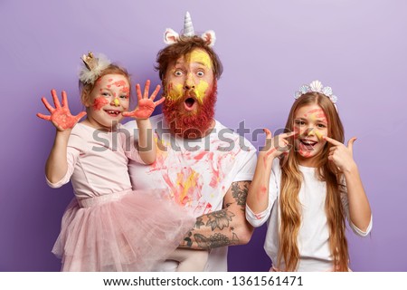 Small female child shows hands soiled with colourful gouache paintings, her glad sister has stains of watercolour on face, stunned father stares at camera, have fun before mother returns from work.