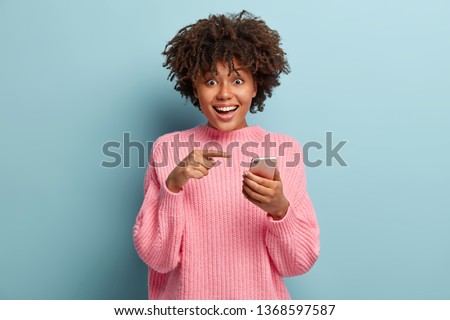 Surprised positive dark skinned lady points at smart phone device, suggests to have conversation in group chat, glad to recieve many messages of congratulation on mailbox, has overjoyed look
