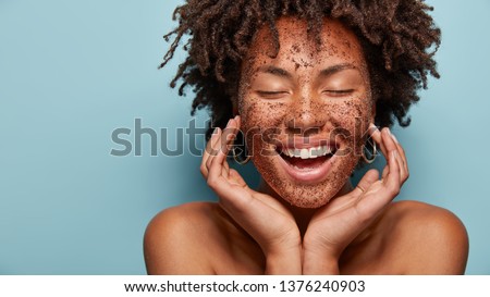Cheerful dark skinned lady applies coffee scrub on face, pampers skin, closes eyes from pleasure, smiles positively has bare shoulders, stands against blue background, free space aside. Beauty concept
