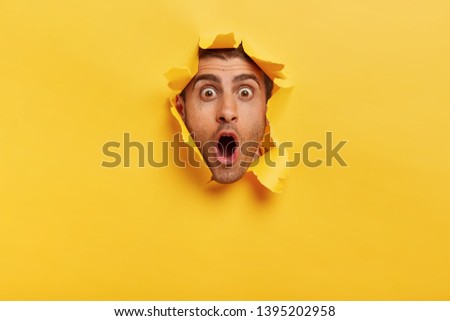 Stupefied young man with bristle keeps jaw dropped from surprisement, has popped eyes, stares through yellow torn paper, impressed by shocking rumors. People, human emotions and reaction concept Foto d'archivio © 